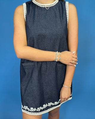 A sleeveless dark blue denim dress with white floral lace trim at the neckline and hem, shown on a model against a solid blue background.