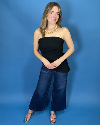 A woman modeling a black strapless bandeau top and dark blue wide-leg jeans against a solid blue studio backdrop.