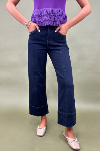 Person wearing a purple top with fringe and dark blue jeans on a plain background