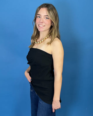 A woman modeling a black strapless top with a ruched front, paired with blue jeans and a gold beaded necklace