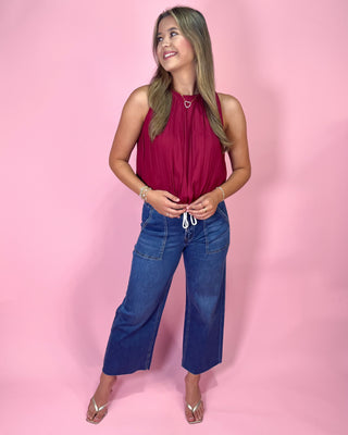 Woman wearing a red sleeveless top and blue jeans against a pink background