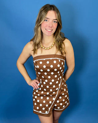 A woman modeling a brown and white polka dot strapless romper against a solid blue background.