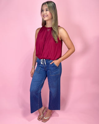 Woman wearing a red sleeveless top and blue jeans against a pink background