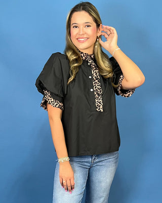 A woman modeling a black puff-sleeve blouse with leopard-print trim on the collar and cuffs, paired with light-wash blue jeans