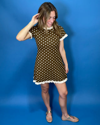 A woman modeling a brown short-sleeve mini dress with white polka dots and white ruffle trim