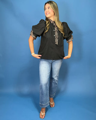 A woman modeling a black puff-sleeve blouse with leopard-print trim on the collar and cuffs, paired with light-wash blue jeans