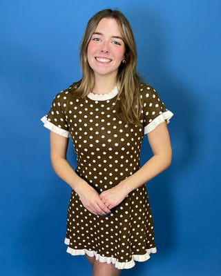 A young woman smiling and wearing a brown short-sleeve dress with white polka dots and white ruffled trim