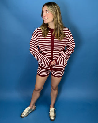 A woman modeling a matching red and white striped knit lounge set consisting of a long-sleeve top and shorts, paired with silver sneakers