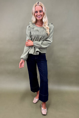 Woman wearing a green blouse and dark jeans against a plain background