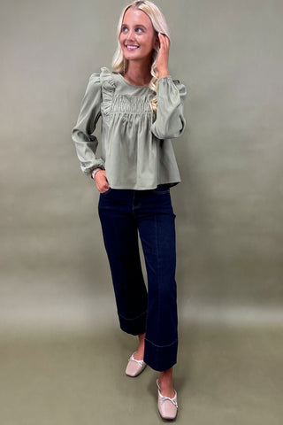 Woman wearing a green blouse and dark jeans against a plain background