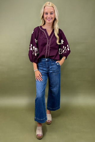 Woman wearing a purple blouse with floral embroidery and blue jeans on a green background