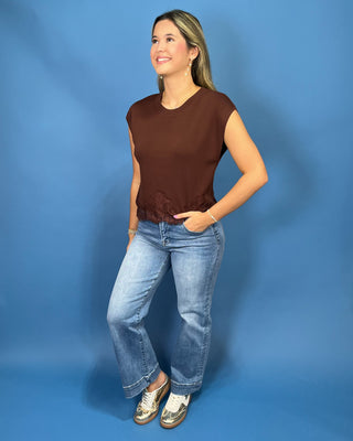 A woman modeling a brown sleeveless top with a ruffled hem, paired with light-wash blue jeans and white sneakers