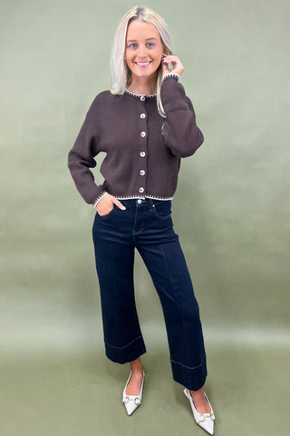 Woman wearing a brown cardigan and dark blue jeans against a green background