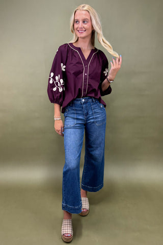 Woman wearing a purple blouse with floral embroidery and blue jeans on a plain background