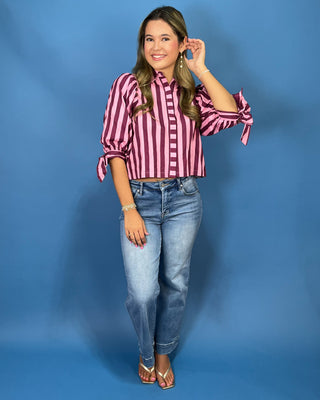 A woman modeling a pink and burgundy striped button-up blouse with voluminous sleeves and tie details at the cuffs