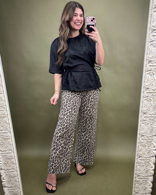 Woman taking a mirror selfie wearing a black top and leopard print pants on a green background.