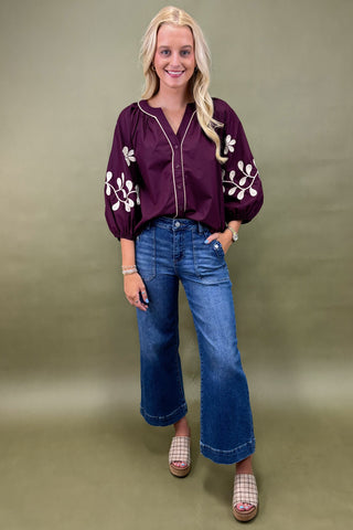 Woman wearing a purple blouse with white floral embroidery and blue jeans on a green background