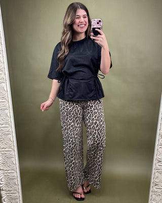 Woman taking a mirror selfie wearing a black top and leopard print pants on a green background