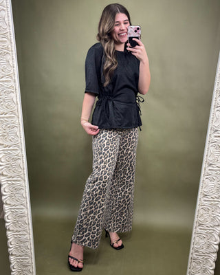 Woman taking a mirror selfie wearing a black top and leopard print pants on a green background.