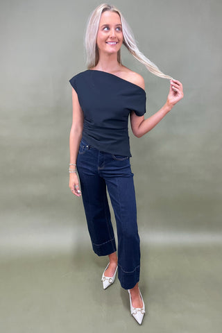 Woman wearing a black one-shoulder top and dark jeans on a plain background