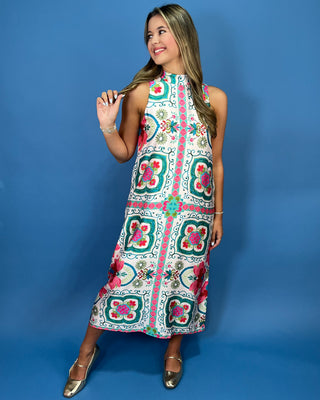 A woman modeling a sleeveless midi dress with a colorful geometric and floral pattern, standing against a solid blue studio backdrop.