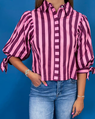 A woman modeling a pink and burgundy striped button-up blouse with voluminous sleeves and tie details at the cuffs