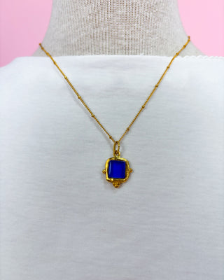 Gold necklace with a blue pendant on a mannequin against a pink background