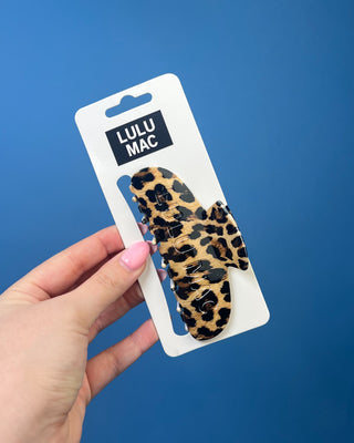 A leopard print hair claw clip in its white retail packaging, held by a hand against a solid blue background. The packaging features the brand name 'LULU MAC' in a black box.