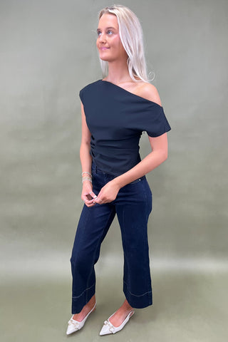 Woman wearing a black one-shoulder top and dark jeans on a plain background