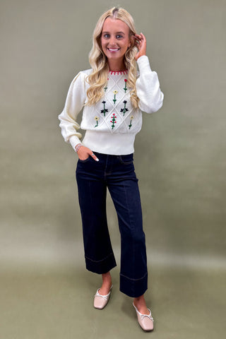 Woman wearing a white sweater with floral patterns and dark blue jeans against a beige background