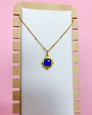 Gold necklace with a blue pendant on a white card, against a pink background