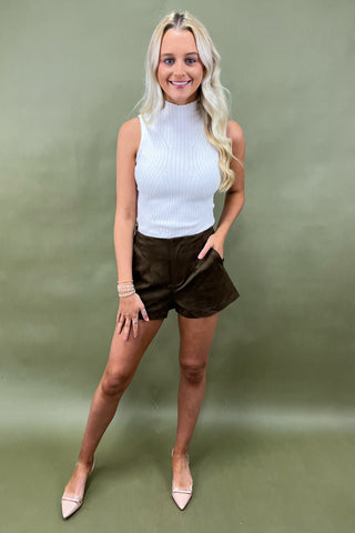 Woman wearing a white sleeveless top and brown shorts against a green background