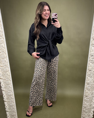 Woman taking a mirror selfie wearing a black top and leopard print pants on a green background.