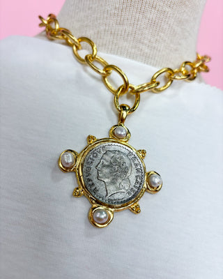 Gold bracelet with a coin and pearl charm on a mannequin arm against a pink background