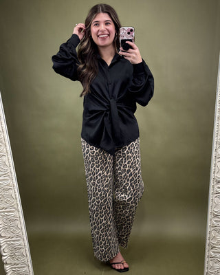 Woman taking a mirror selfie wearing a black top and leopard print pants on a green background