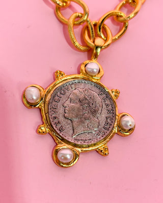 Gold necklace with a vintage coin and pearl pendant on a pink background