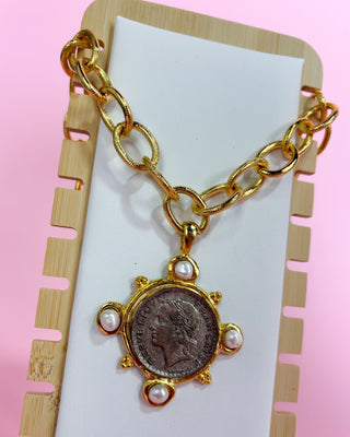 Gold chain necklace with a coin and pearl pendant on a pink background