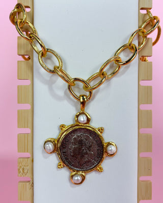 Gold chain necklace with a pendant featuring a coin and pearls on a white card with pink border.