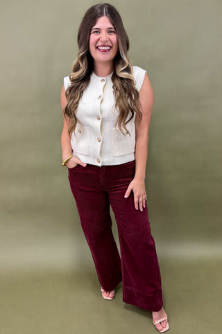 Woman wearing a white sleeveless top and burgundy pants against a green background