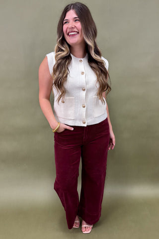 Woman wearing a white sleeveless jacket and burgundy pants against a green background