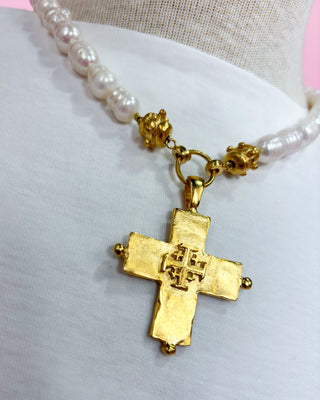 Gold cross pendant on a pearl necklace against a light pink background