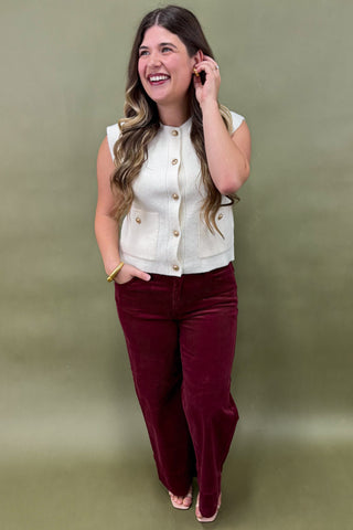 Woman wearing a white blouse with gold buttons and burgundy pants against a green background