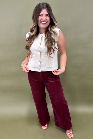Woman wearing a white sleeveless top and burgundy pants against a green background