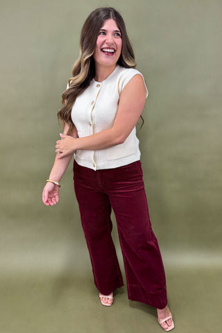 Woman wearing a white sleeveless top and burgundy pants against a green background