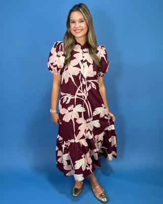 A woman modeling a short puff-sleeve midi dress with a burgundy and cream floral pattern, standing against a solid blue studio background.