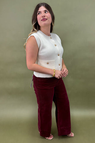 Woman wearing a white sleeveless top with gold buttons and burgundy pants against a green background