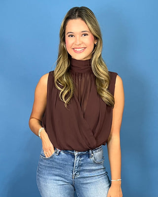 A woman with blonde-highlighted hair wearing a sleeveless, high-neck brown blouse and blue jeans