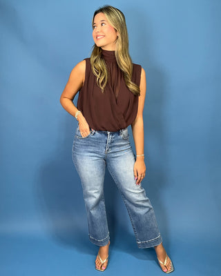 A woman modeling a sleeveless, high-neck brown top paired with light-wash blue jeans and metallic sandals