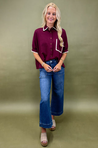 Woman wearing a burgundy shirt with white trim and blue jeans against a green background