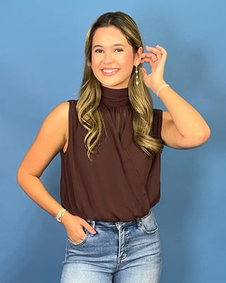 A woman with blonde-highlighted hair wearing a sleeveless high-neck brown top and blue jeans